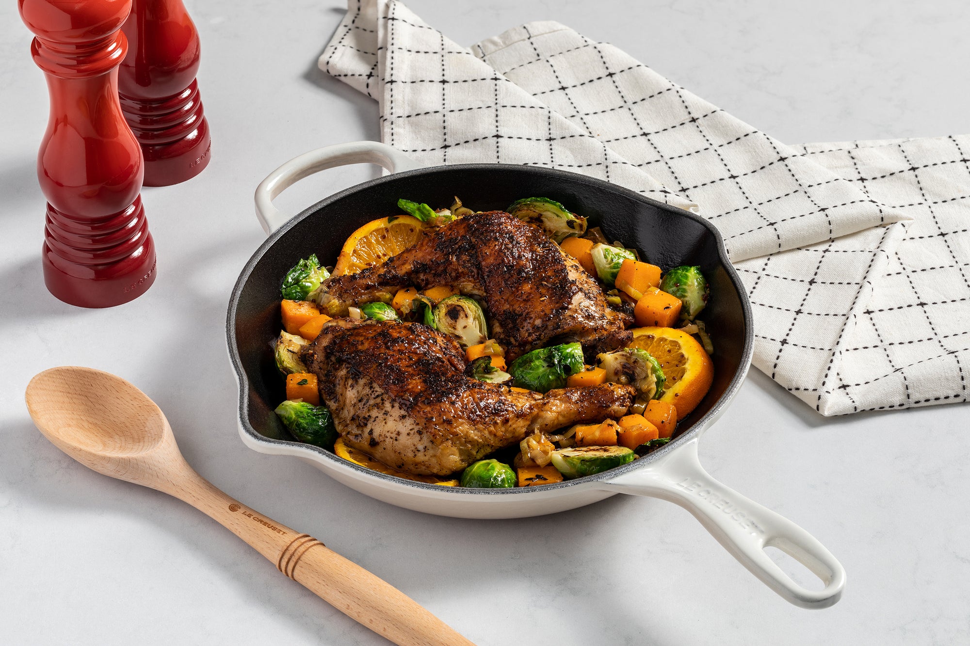 A Le Creuset Signature Skillet full of roast chicken, veggies, and oranges sits on a marble countertop alongside other kitchen tools.