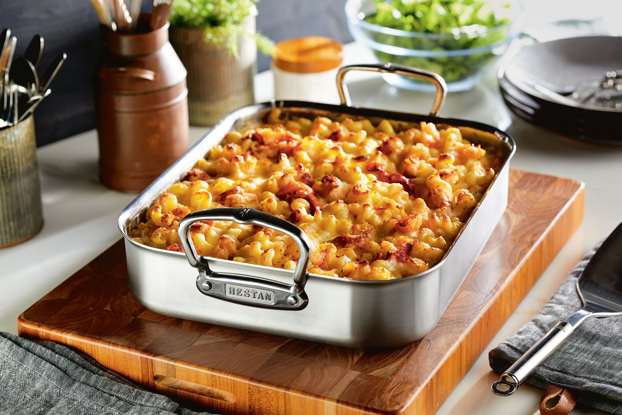 A Hestan roasting pan filled with baked pasta sits on a wooden cutting board.