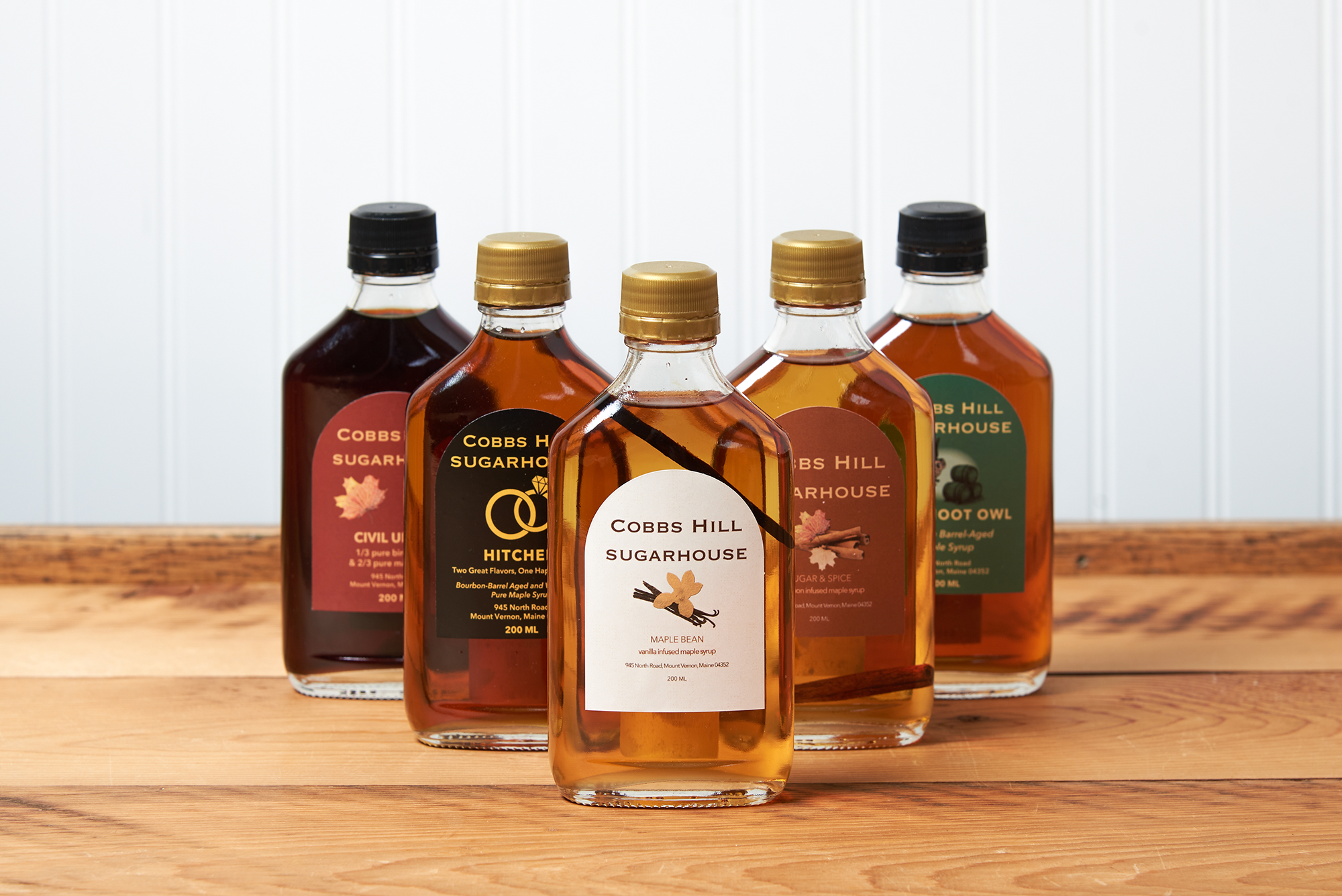 Five bottles of Cobb's Hill Maple Syrup sit on a brown wooden table. The flavors include: Maple Bean, Sugar and Spice, Tipsy Hoot Owl, Civil Union, and Hitched.