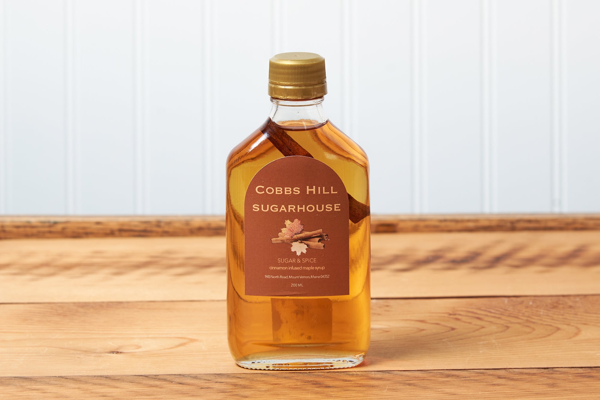 A bottle of Cobb's Hill Sugar and Spice Maple Syrup sits on a brown wooden table.