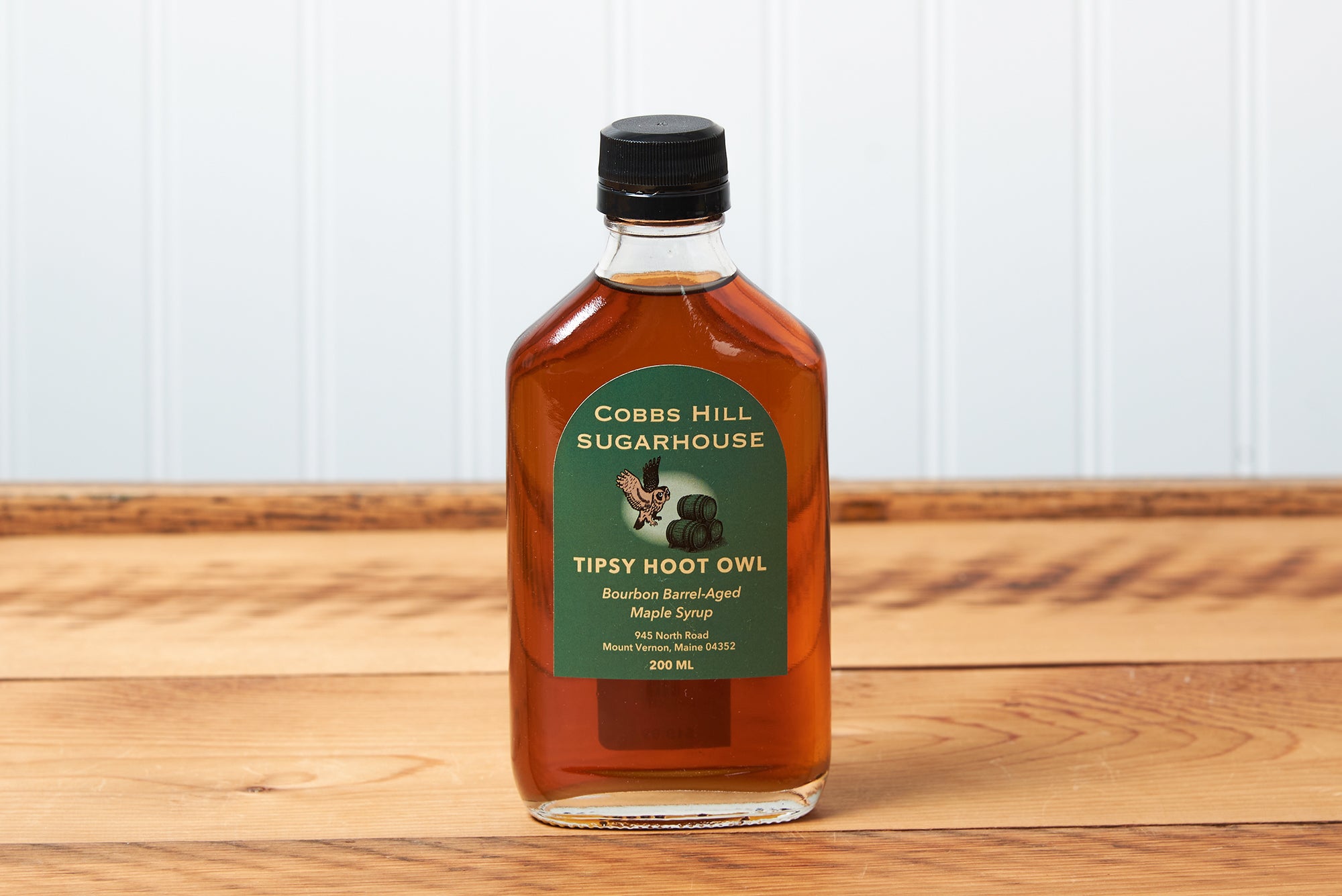 A bottle of Cobb's Hill Tipsy Hoot Owl Maple Syrup sits on a brown wooden table.