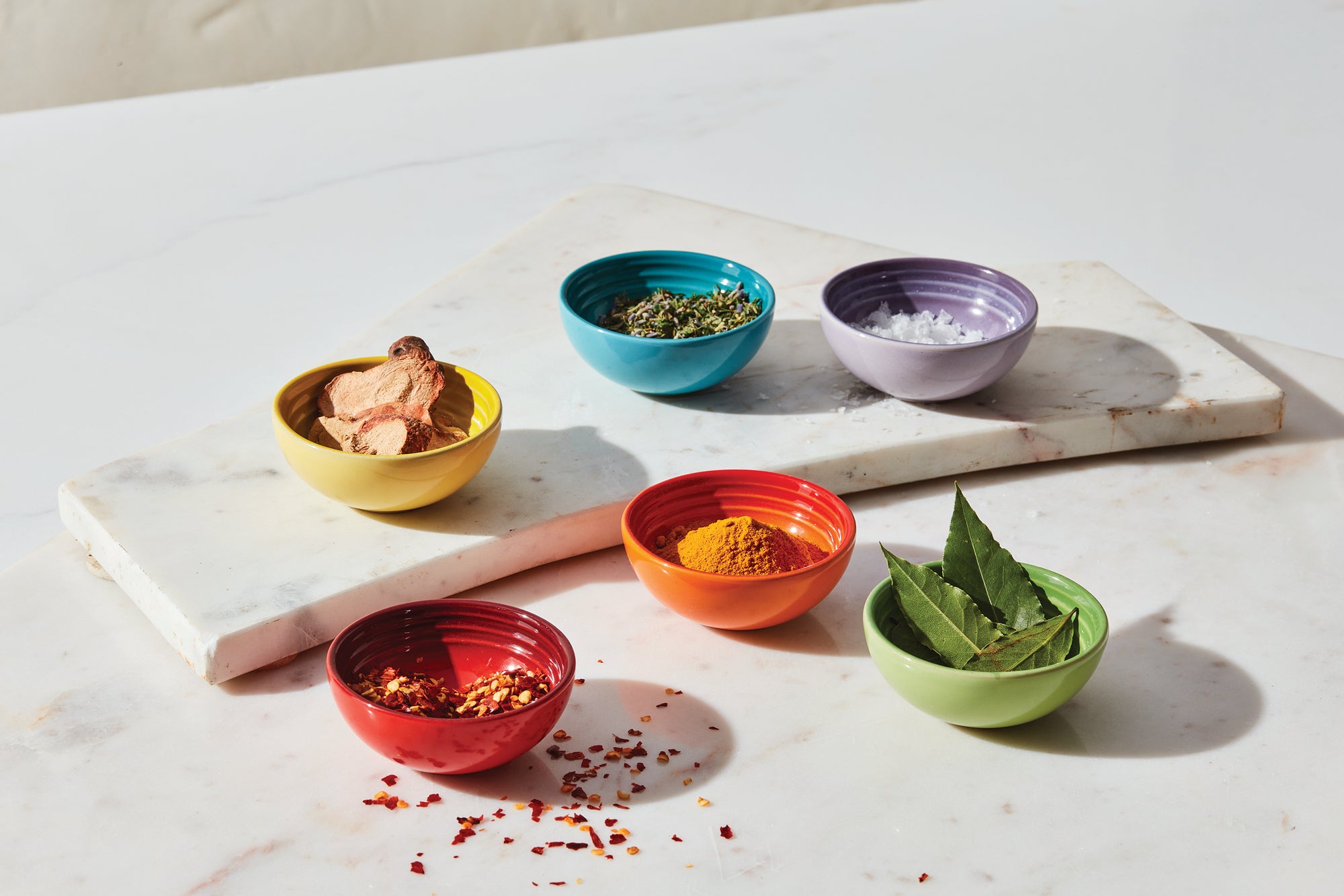 Six Le Creuset Pinch Bowls in assorted colors sit on a white marble countertop. Each bowl holds ingredients, including spices and herbs.