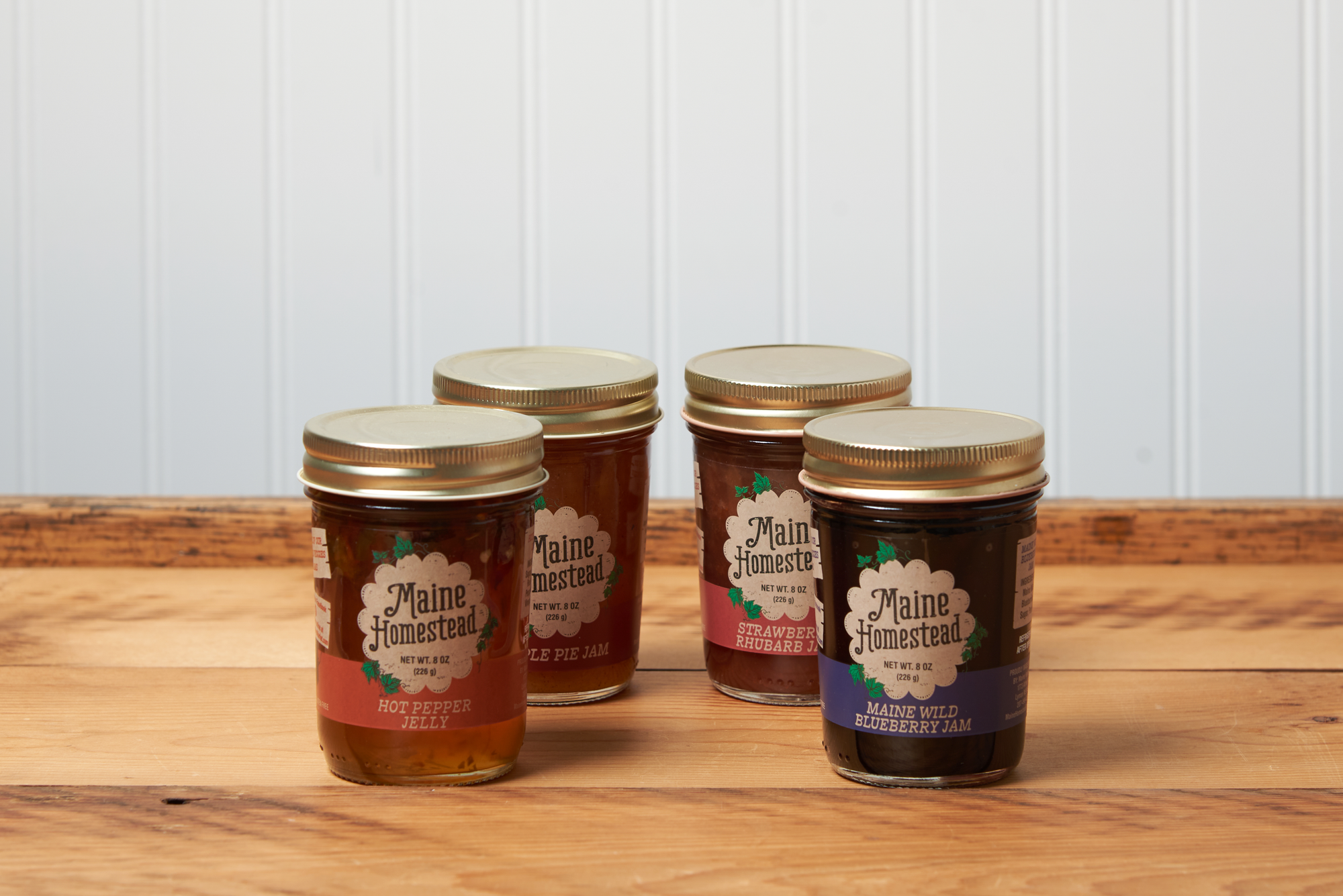 Four jars of assorted Maine Homestead jams and jelly sit on a brown wooden table.