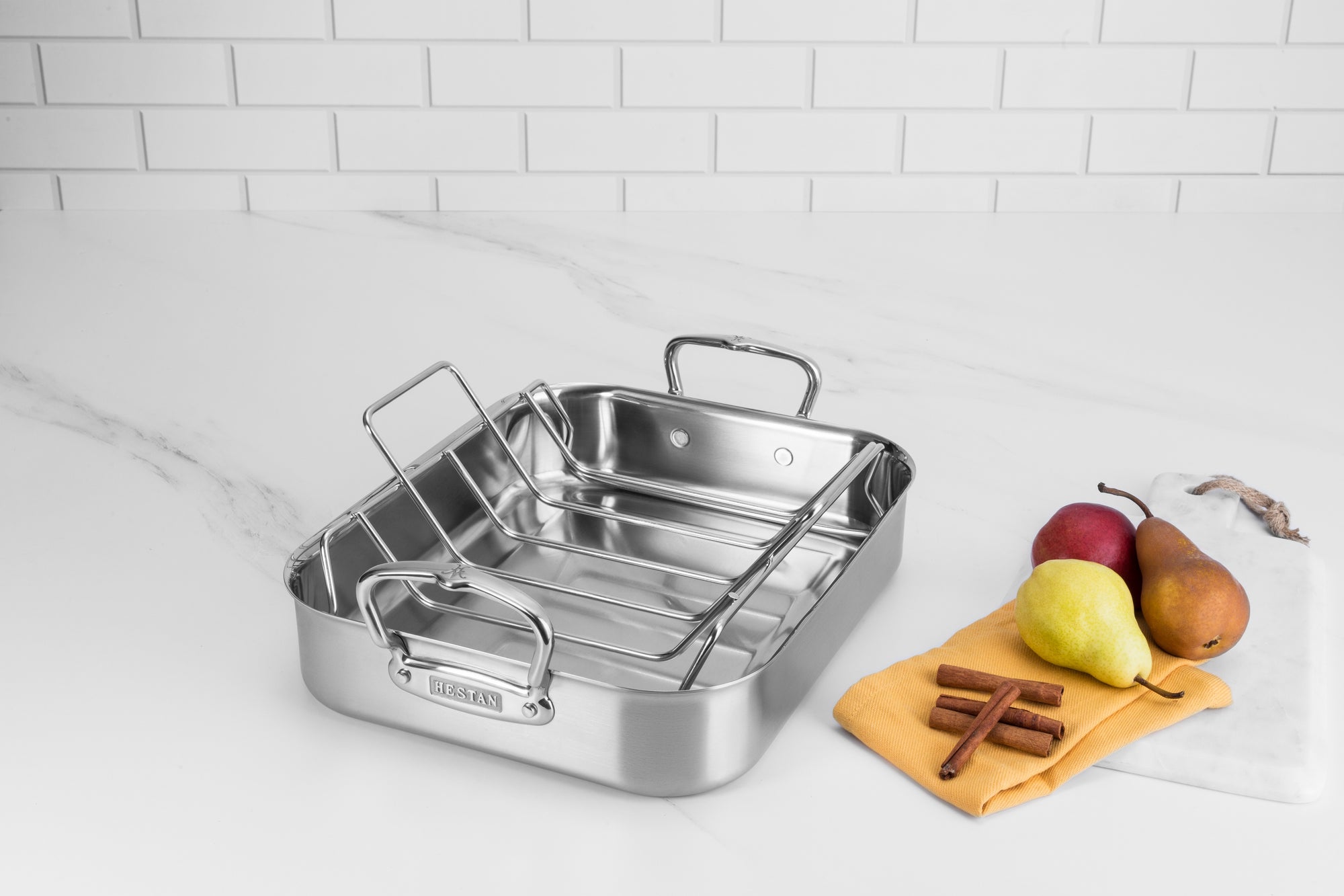 A small Hestan roasting pan sits on a white marble countertop alongside pears, cinnamon sticks, and a yellow kitchen towel.