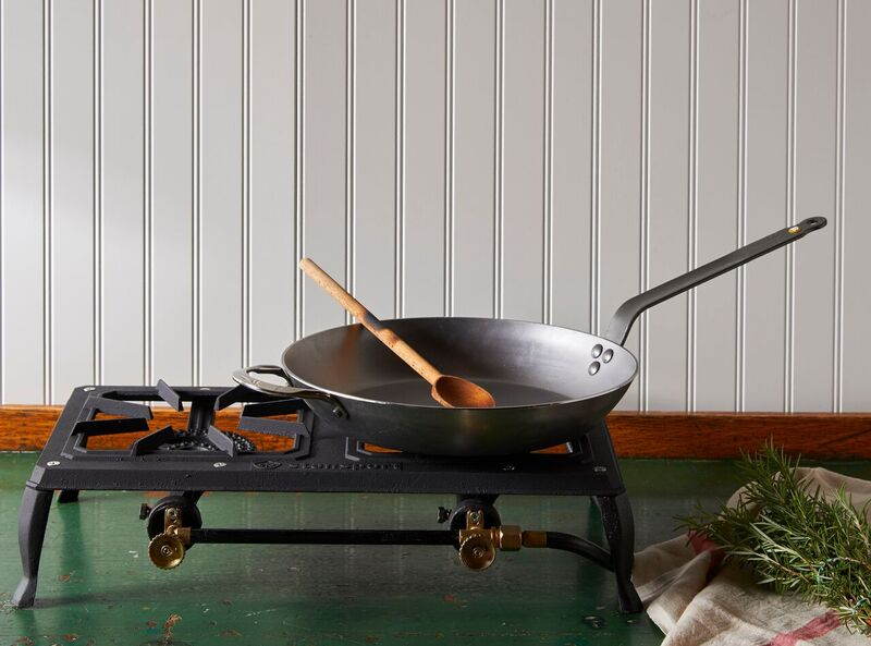A Debuyer Mineral B Carbon Steel Fry Pan with a wooden spoon in it rests on a portable stovetop, which sits on a green table in front of a white wall.