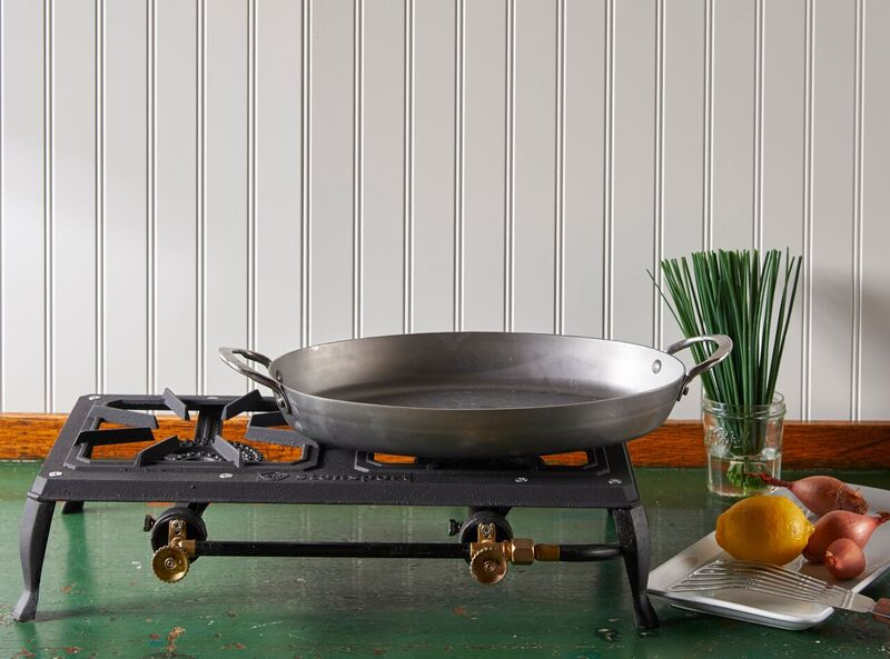 A Mineral B Carbon Steel Oval Fry Pan rests on a portable stovetop, which sits on a green table in front of a white wall.