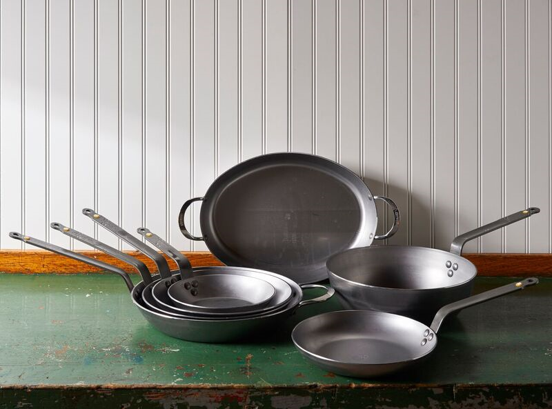 A collection of Debuyer Mineral B Carbon Steel Fry Pans sits on a green table, in front of a white wall.