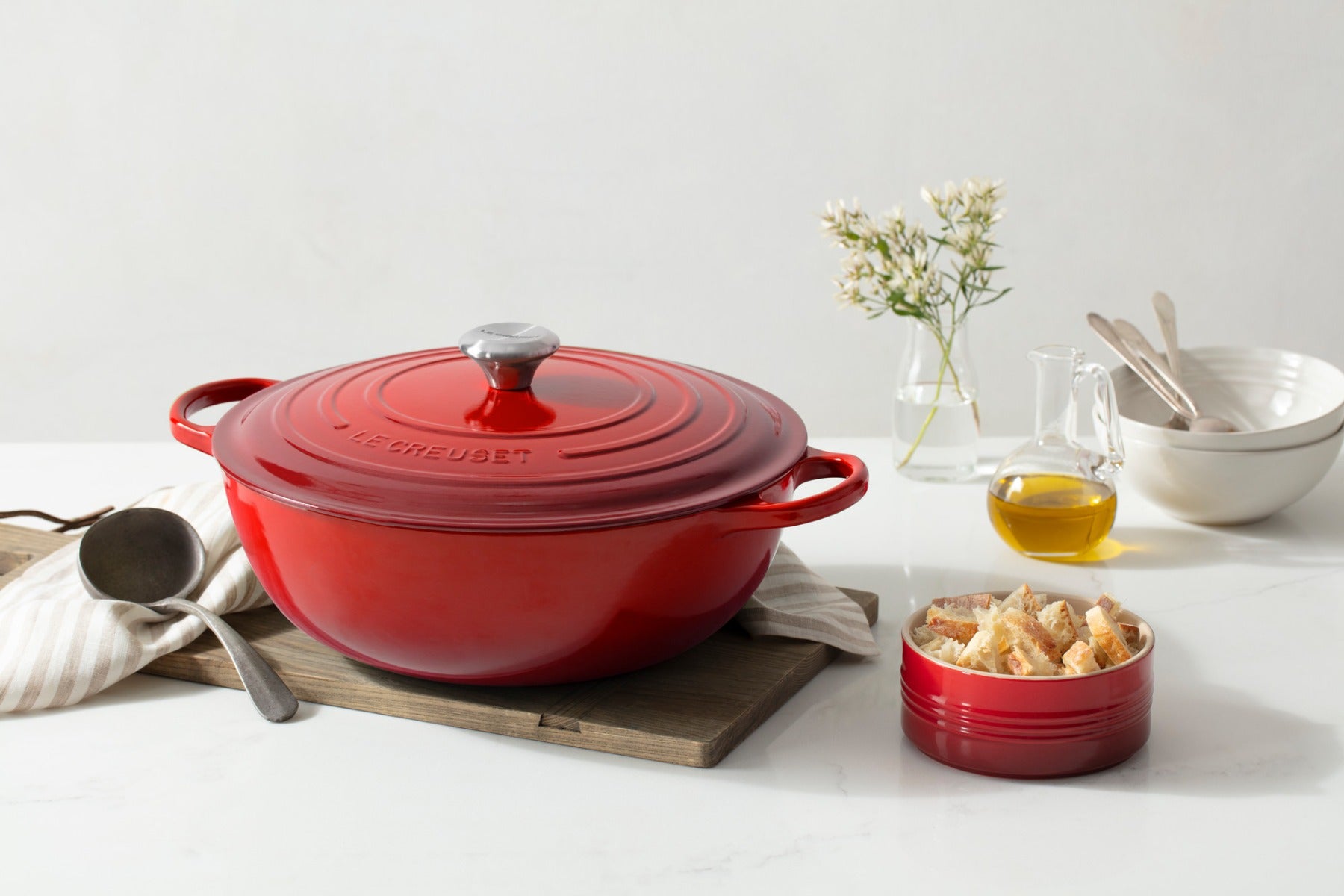 Red enameled cast iron Dutch oven on wooden board with small bread bowl and olive oil.