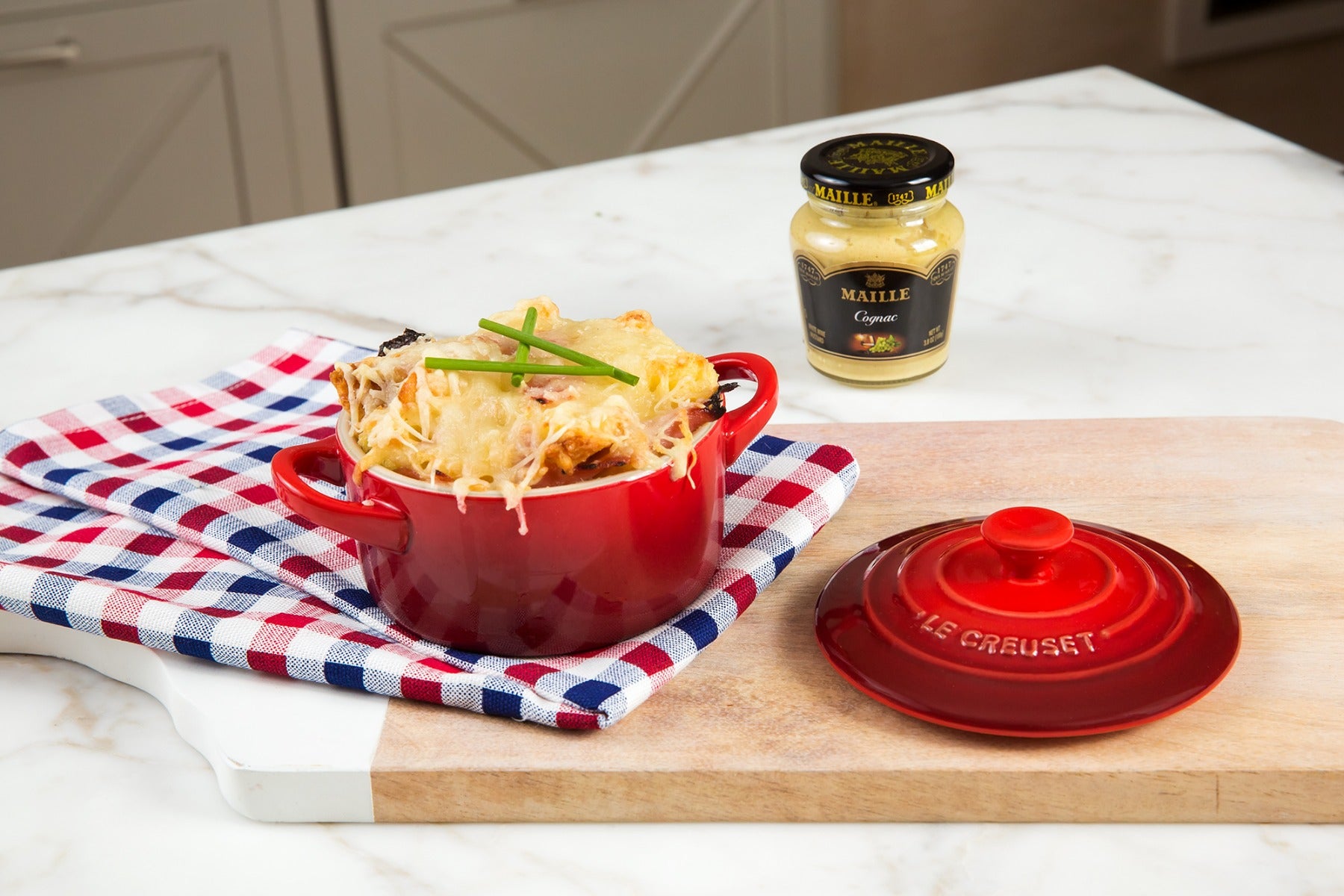 Cheesy baked dish in red Le Creuset pot next to Maille mustard jar
