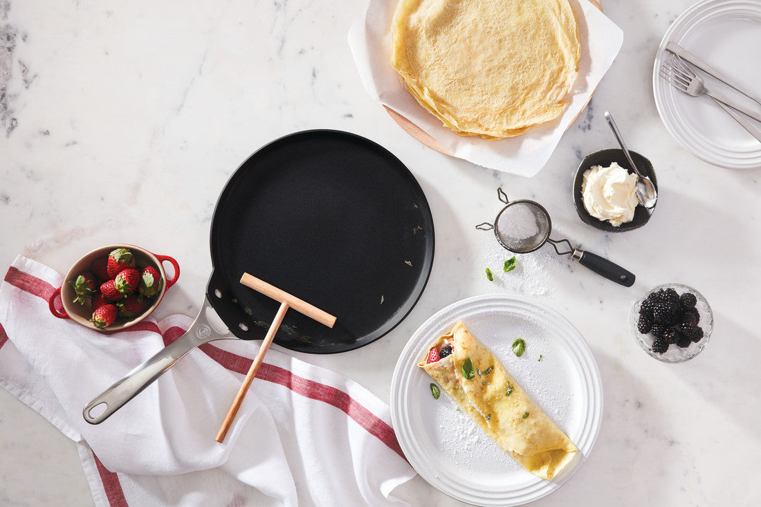 Crepe making ingredients on marble surface with skillet, fresh berries, and folded crepe.