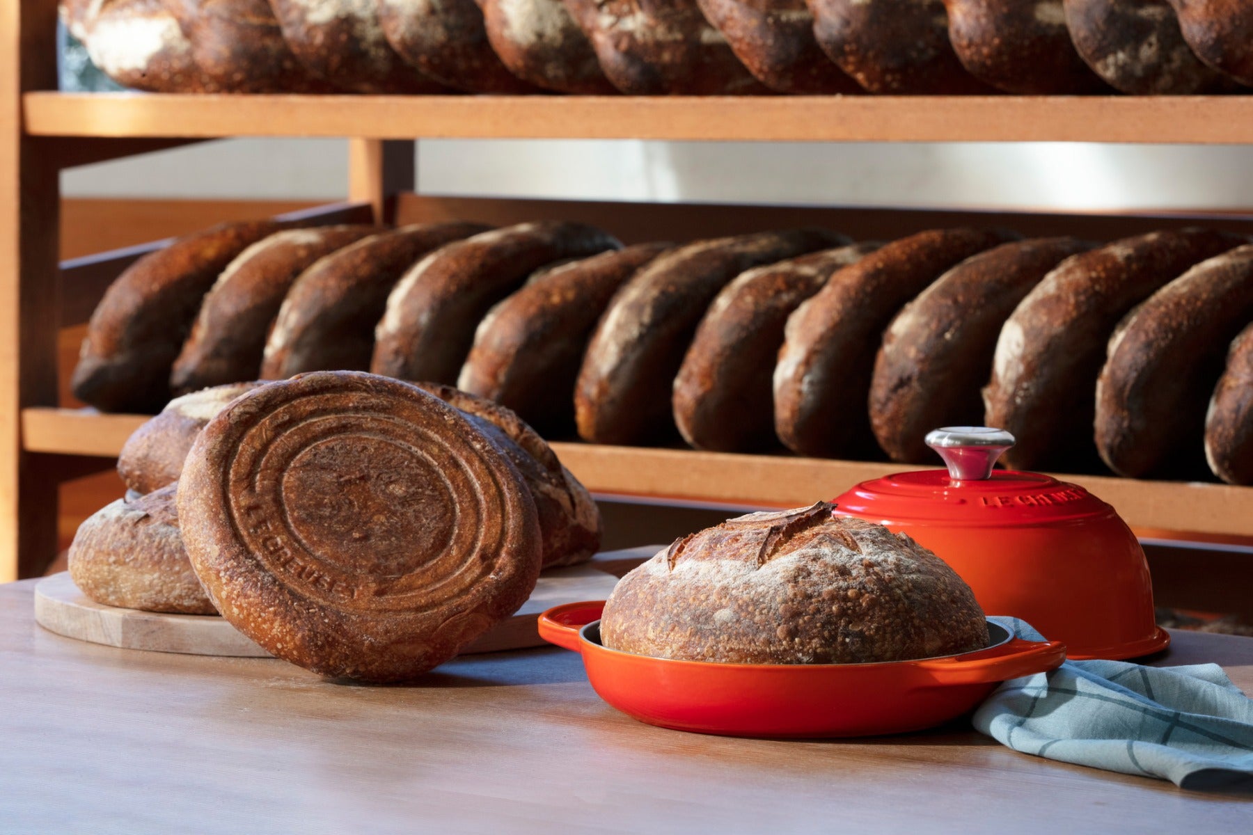 Fresh artisan bread on cutting board with red ceramic bakeware and rack of loaves in background