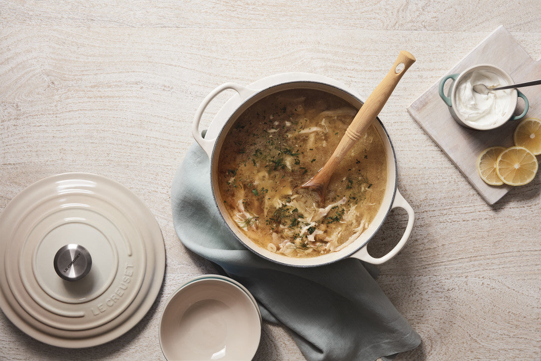 Creamy chicken soup in white pot with wooden spoon, sour cream, and lemon slices on side.