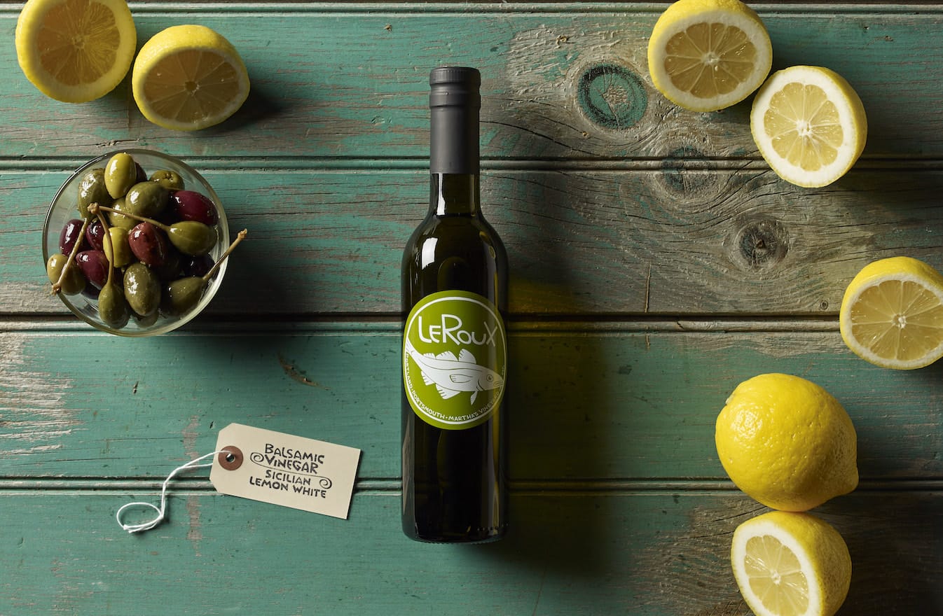 Bottle of LeRoux vinegar, bowl of olives, and lemons on a teal wooden table.
