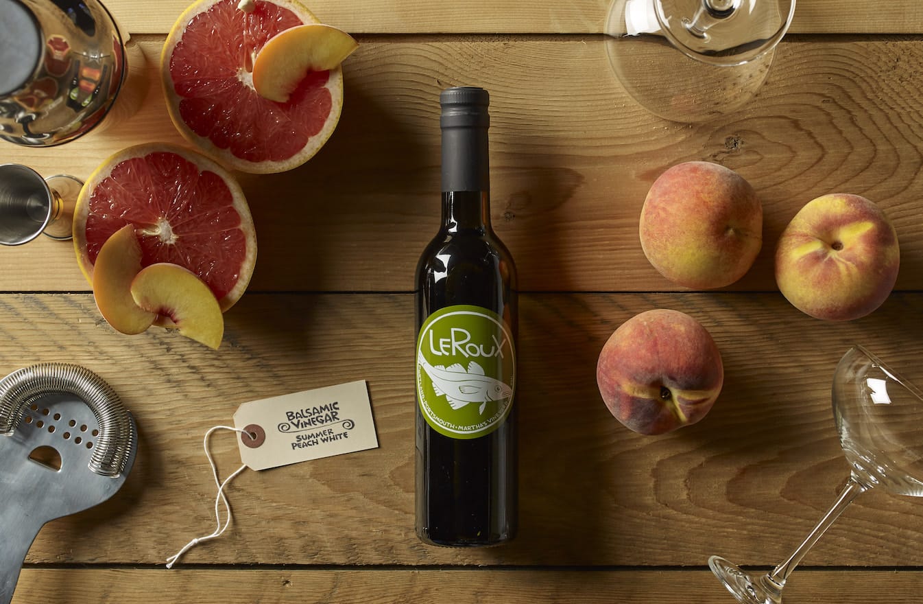 Bottle of LeRoux balsamic vinegar with fresh peaches and grapefruit on a wooden table.