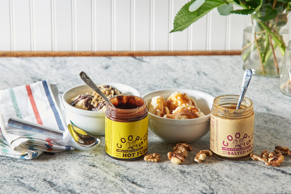 Bowls of ice cream sit on a marble countertop alongside open jars of fudge and caramel.  