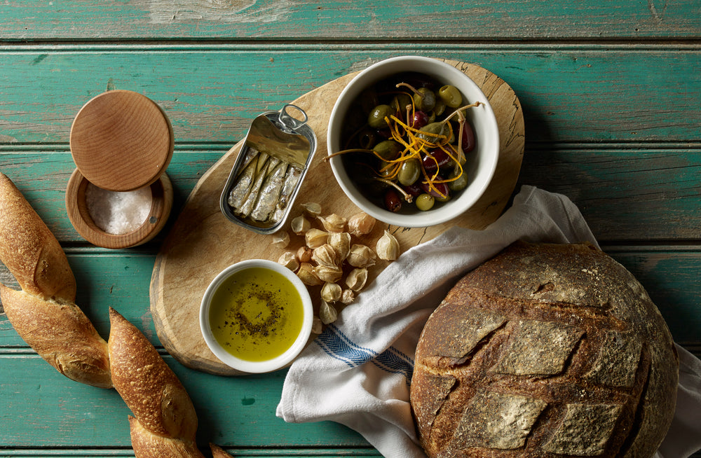 A ramekin filled with olive oil sits alongside olives, sardines, and garlic on a wooden cutting board. The cutting board is on a turquoise table, surrounded by loaves of bread and a salt cellar.