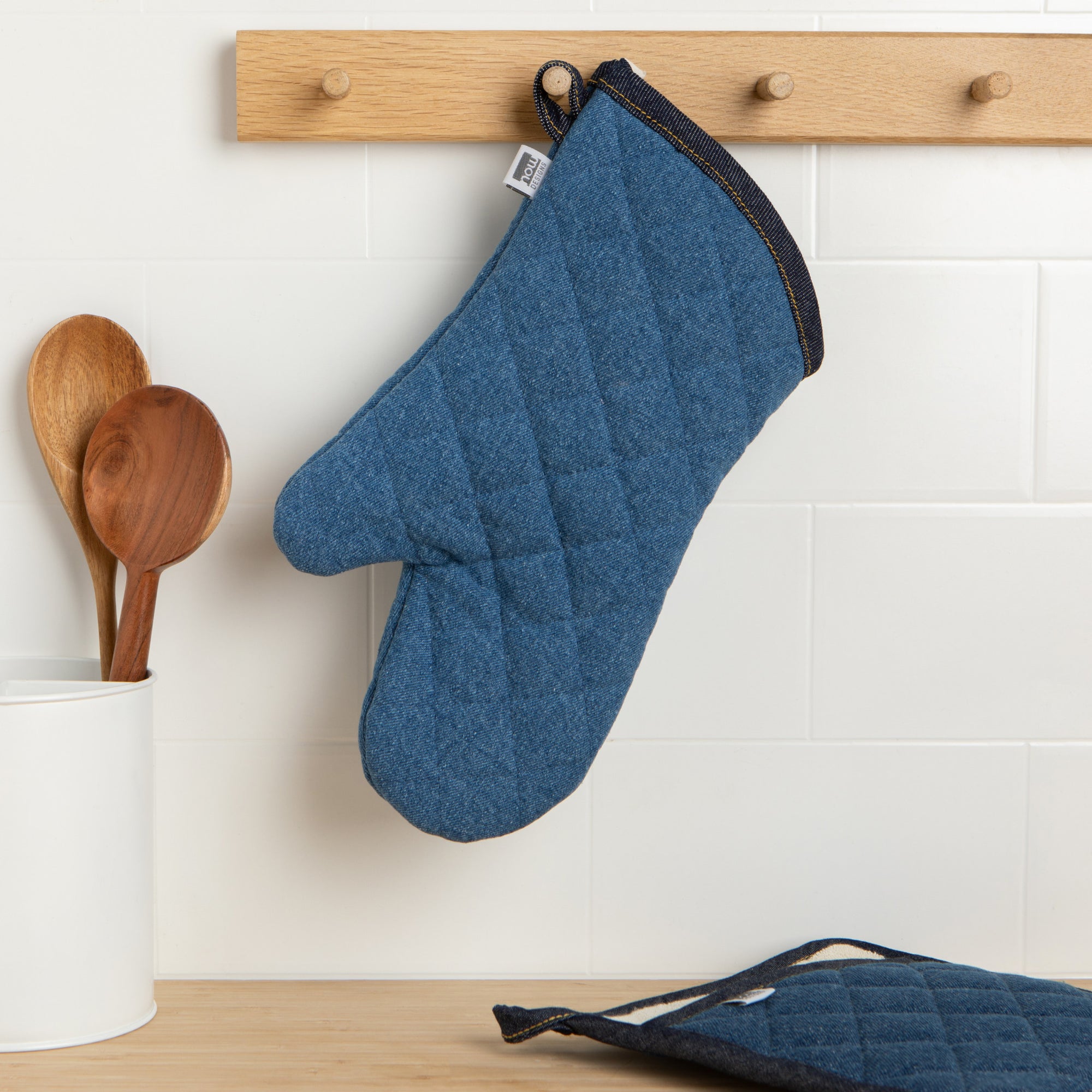 A Now Designs Denim Oven Mitt hangs on a kitchen wall.