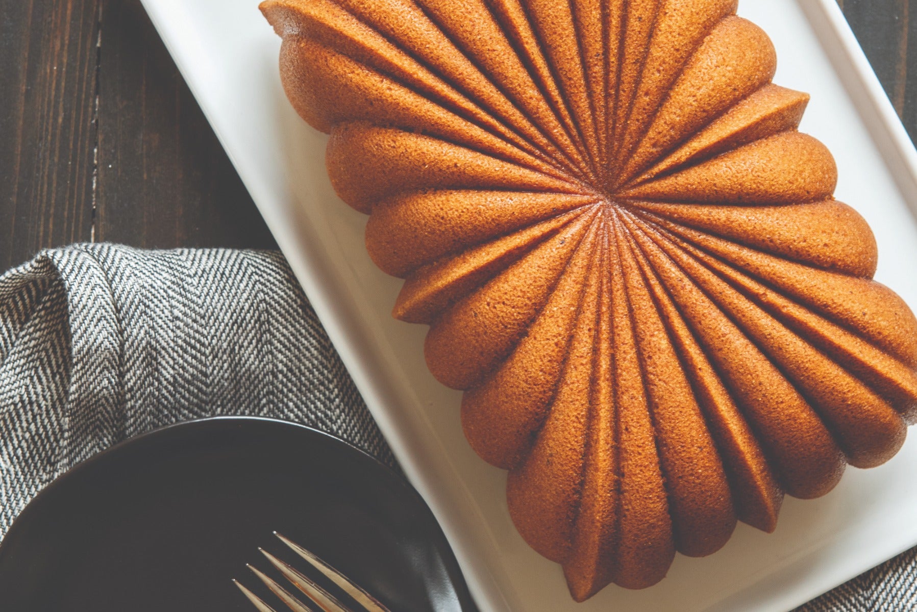 Bronze-colored madeleine cake on white plate with black utensils and napkin