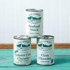 A stack of assorted Bar Harbor Foods soup cans sits on a turquoise wooden table. The assortment includes: New England Clam Chowder, Lobster Corn Chowder, and Seafood Stock.