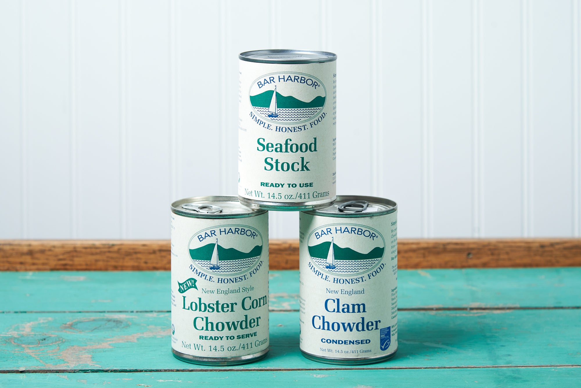 A stack of assorted Bar Harbor Foods soup cans sits on a turquoise wooden table. The assortment includes: New England Clam Chowder, Lobster Corn Chowder, and Seafood Stock.