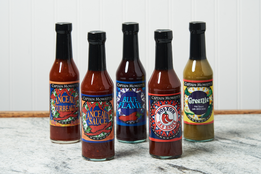 An assortment of Captain Mowatt's Hot Sauce sits on a marble countertop. The flavors include: Blue Flame, Greenie, Canceaux Sauce, Canceaux Turbeaux, and Boston Red Sauce.