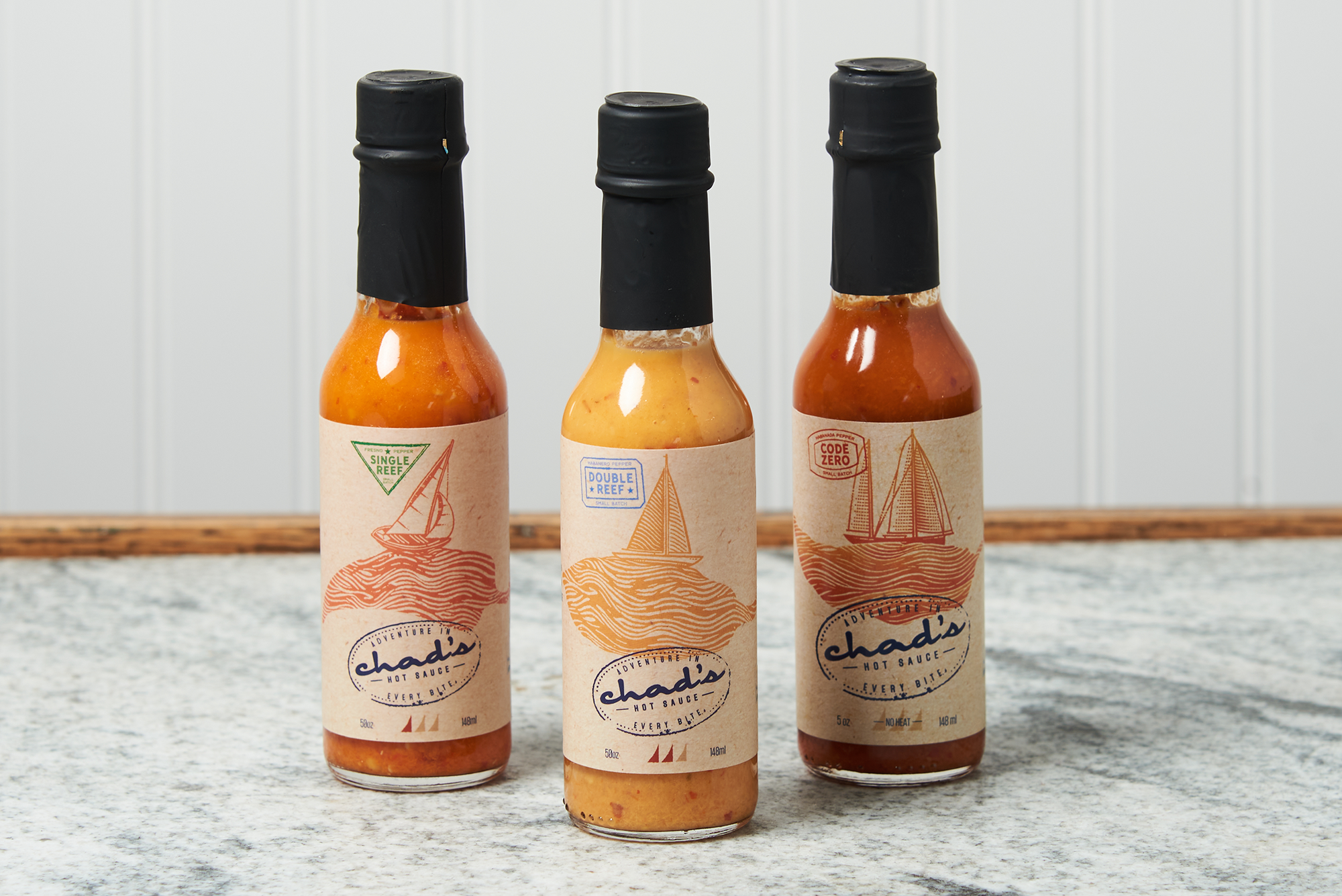 An assortment of Chad's Hot Sauce sits on a marble countertop. The flavors include: Single Reef, Double Reef, and Code Zero - No Heat.