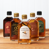 Five bottles of Cobb's Hill Maple Syrup sit on a brown wooden table. The flavors include: Maple Bean, Sugar and Spice, Tipsy Hoot Owl, Civil Union, and Hitched.