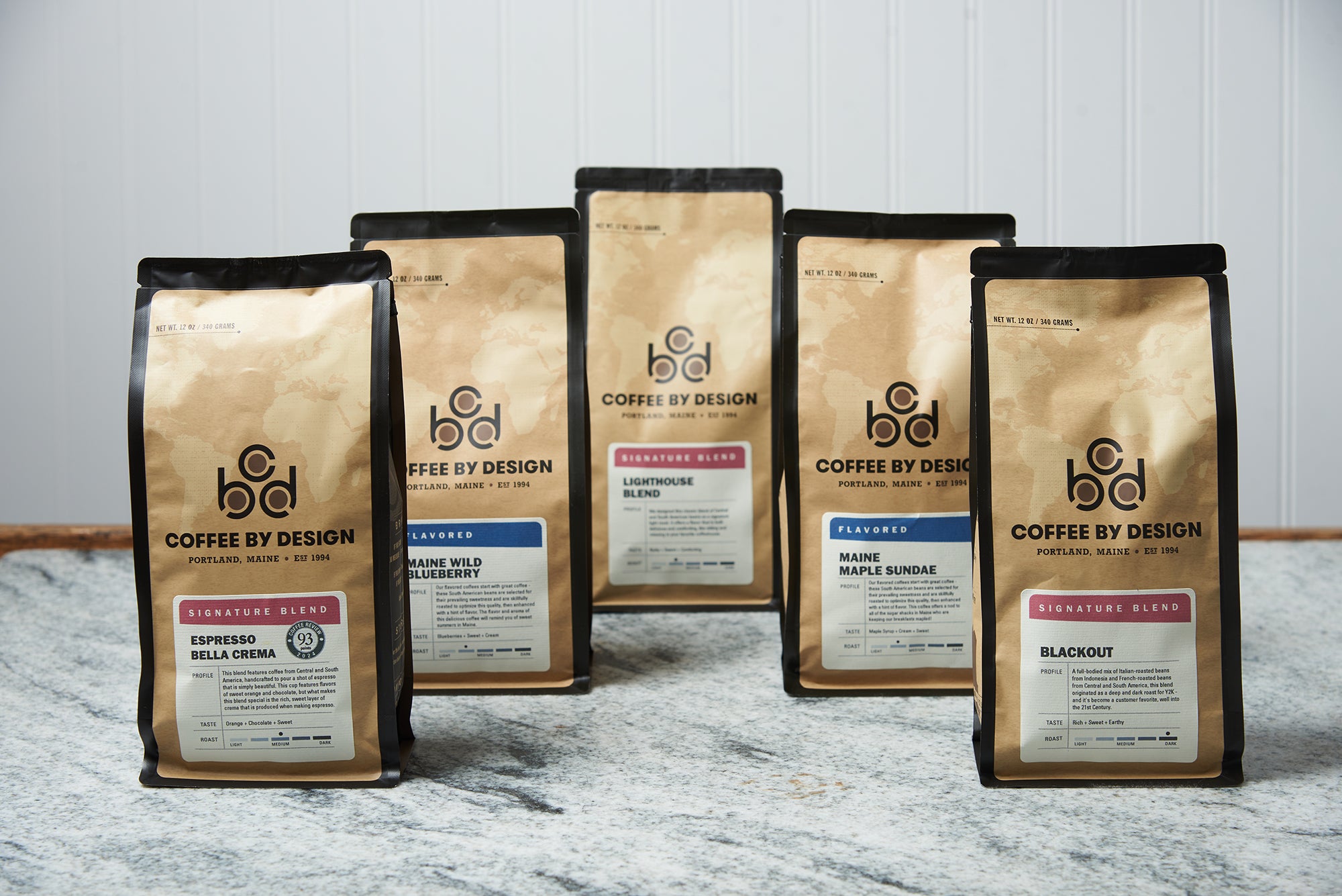 Five bags of Coffee By Design coffee sit on a marble countertop.