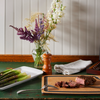 An Epicurean Gourmet Series Cutting Board with Juice Groove sits on a green wooden table. A steak is cut on the board. Beside the board is a box of silver cutlery and a plate of asparagus.