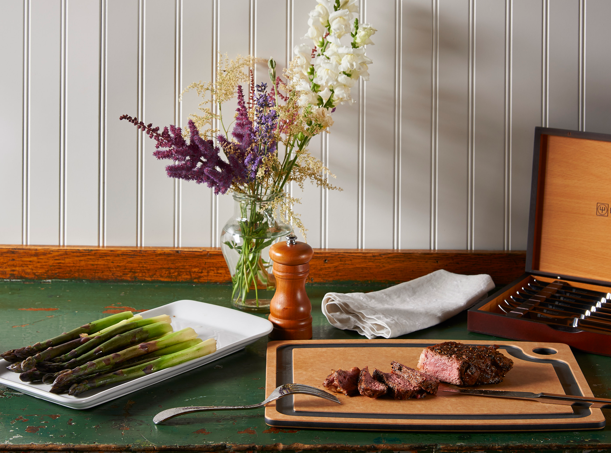 An Epicurean Gourmet Series Cutting Board with Juice Groove sits on a green wooden table. A steak is cut on the board. Beside the board is a box of silver cutlery and a plate of asparagus.