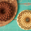 A pumpkin bundt cake sits on a cutting board. Alongside the cake, the bundt pan in which the cake was baked sits on a turquoise wooden table.