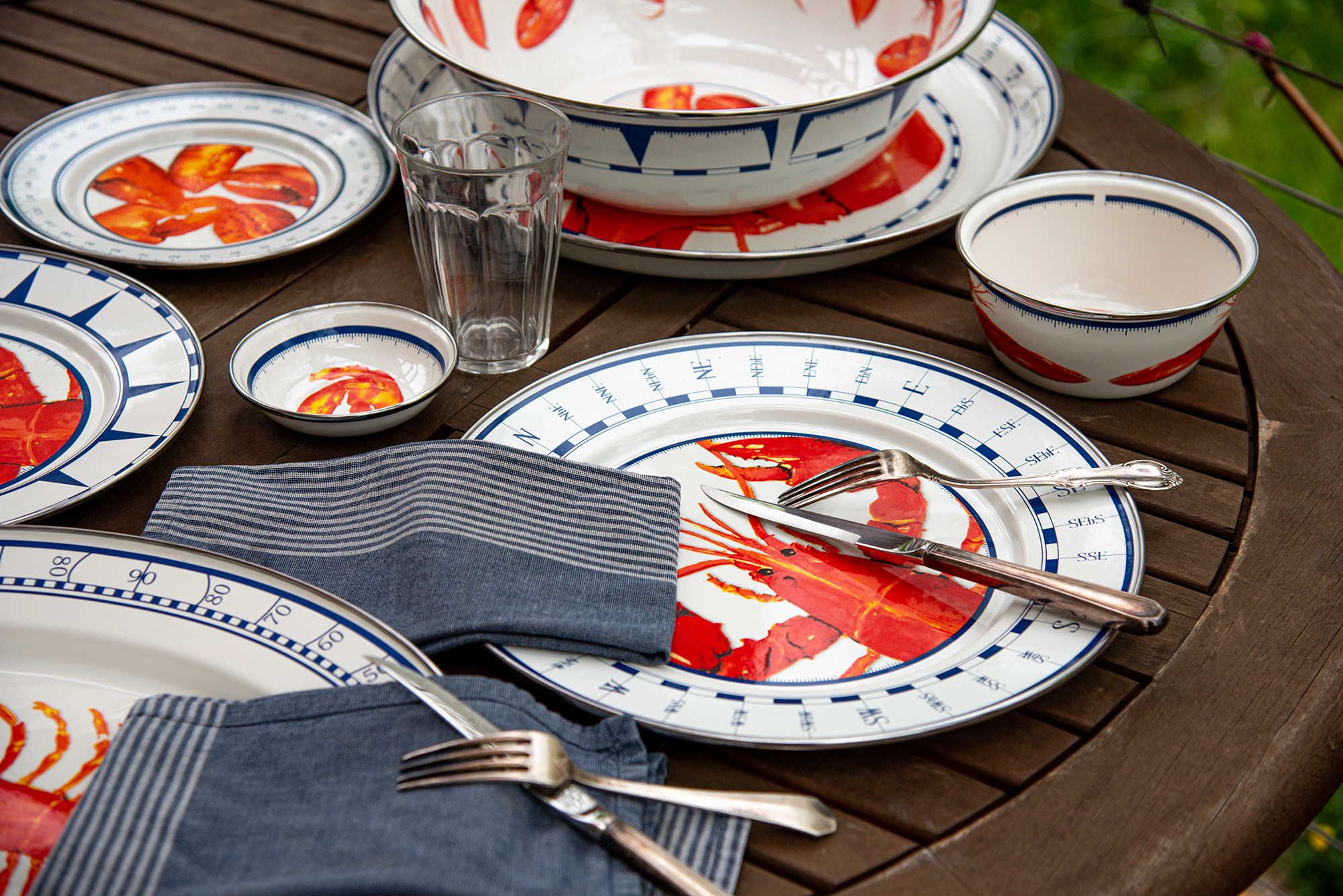 Golden Rabbit Enamelware, decorated with lobster and nautical motifs, sits on a picnic table with silverware, table linens, and an empty glass.