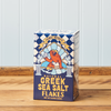A front-facing view of a box of Greek Sea Salt Flakes sitting on a wooden table.