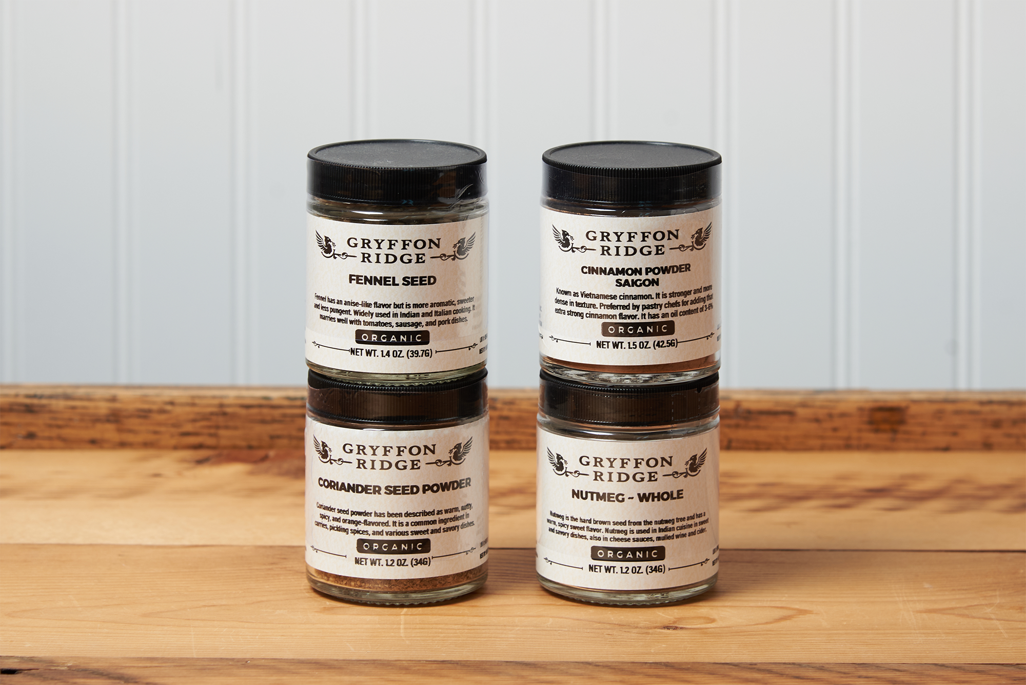 Five jars of Gryffon Ridge spices sit on a wooden tabletop. The spices include Saigon Cinnamon Powder, Coriander Seed Ground, Whole Nutmeg, and Fennel Seed.