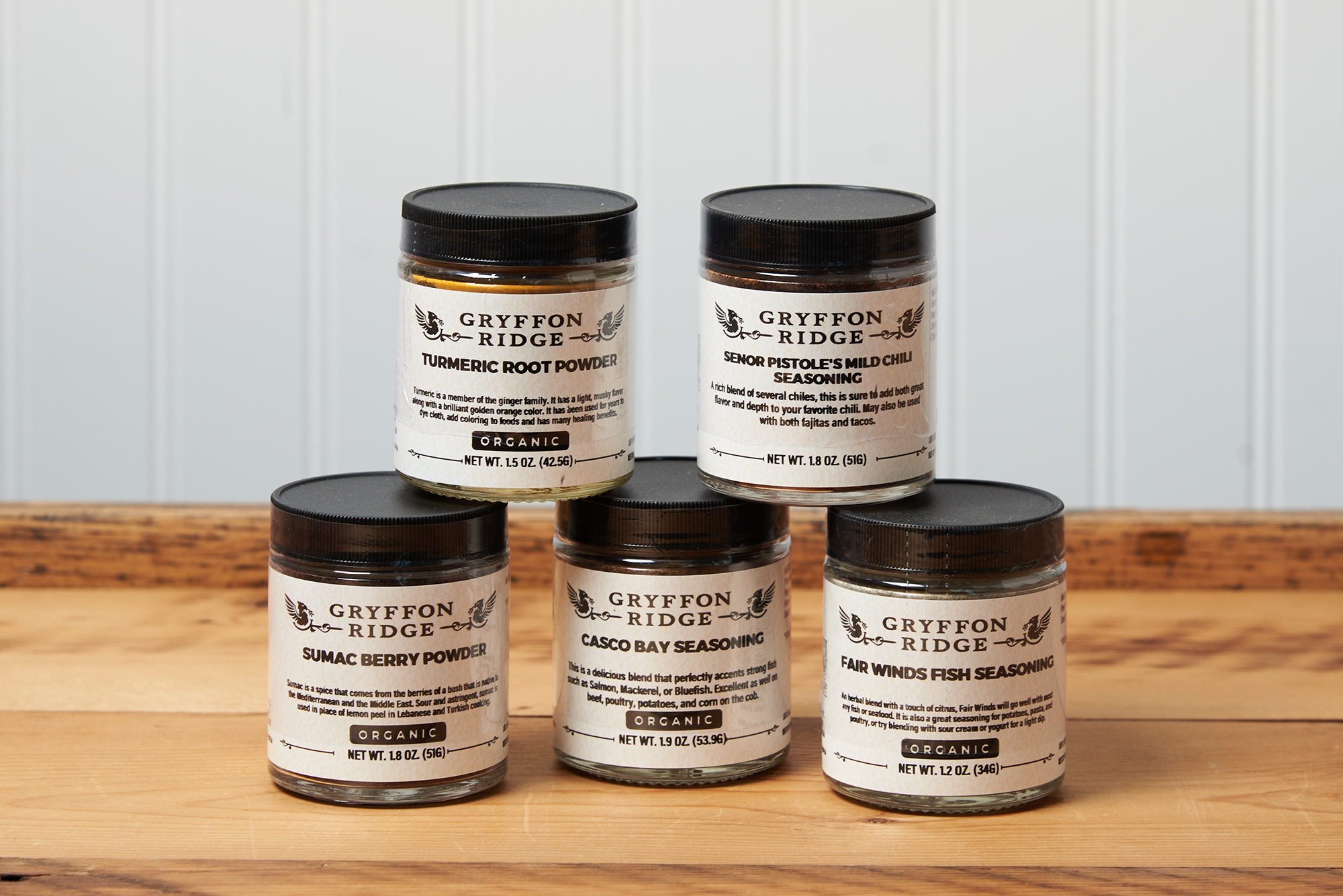 Five jars of Gryffon Ridge spices sit on a wooden tabletop. The spices include Casco Bay Seasoning, Fair Winds Fish Seasoning, Sumac Berry Powder, Turmeric Root Powder, and Senor Pistole’s Mild Chili Seasoning.
