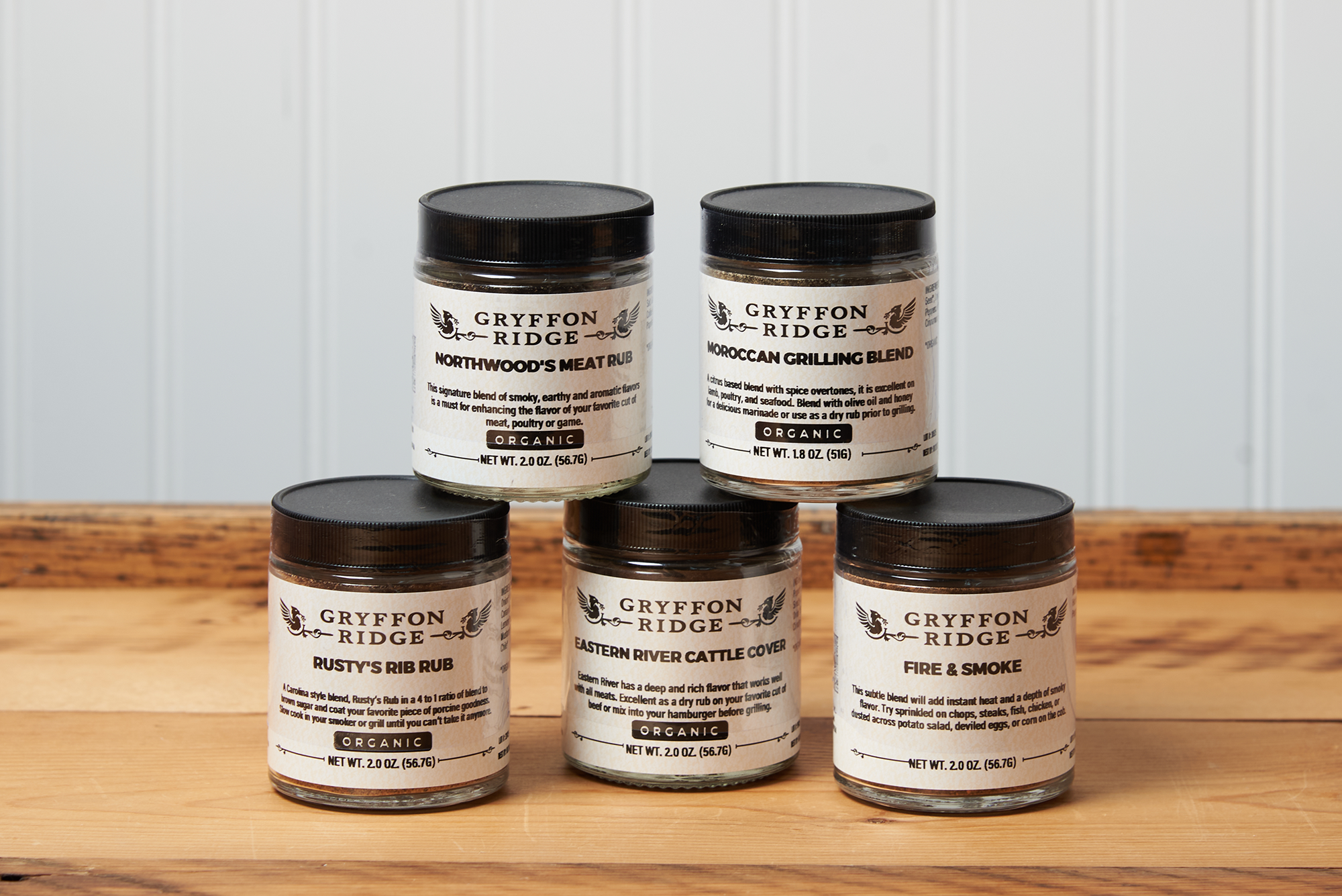 Five jars of Gryffon Ridge spices sit on a wooden tabletop. The spices include Eastern River Cattle Cover, Fire & Smoke, Northwoods Meat Rub, Moroccan Grilling Blend, and Rusty’s Rib Rub.