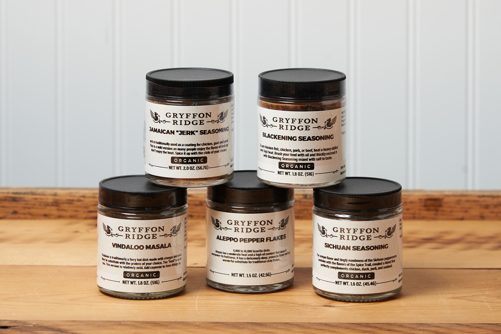 Five jars of Gryffon Ridge spices sit on a wooden tabletop. The spices include Aleppo Pepper Flakes, Blackening Seasoning, Jamaican “Jerk” Seasoning, Vindaloo Masala, and Sichuan Seasoning.