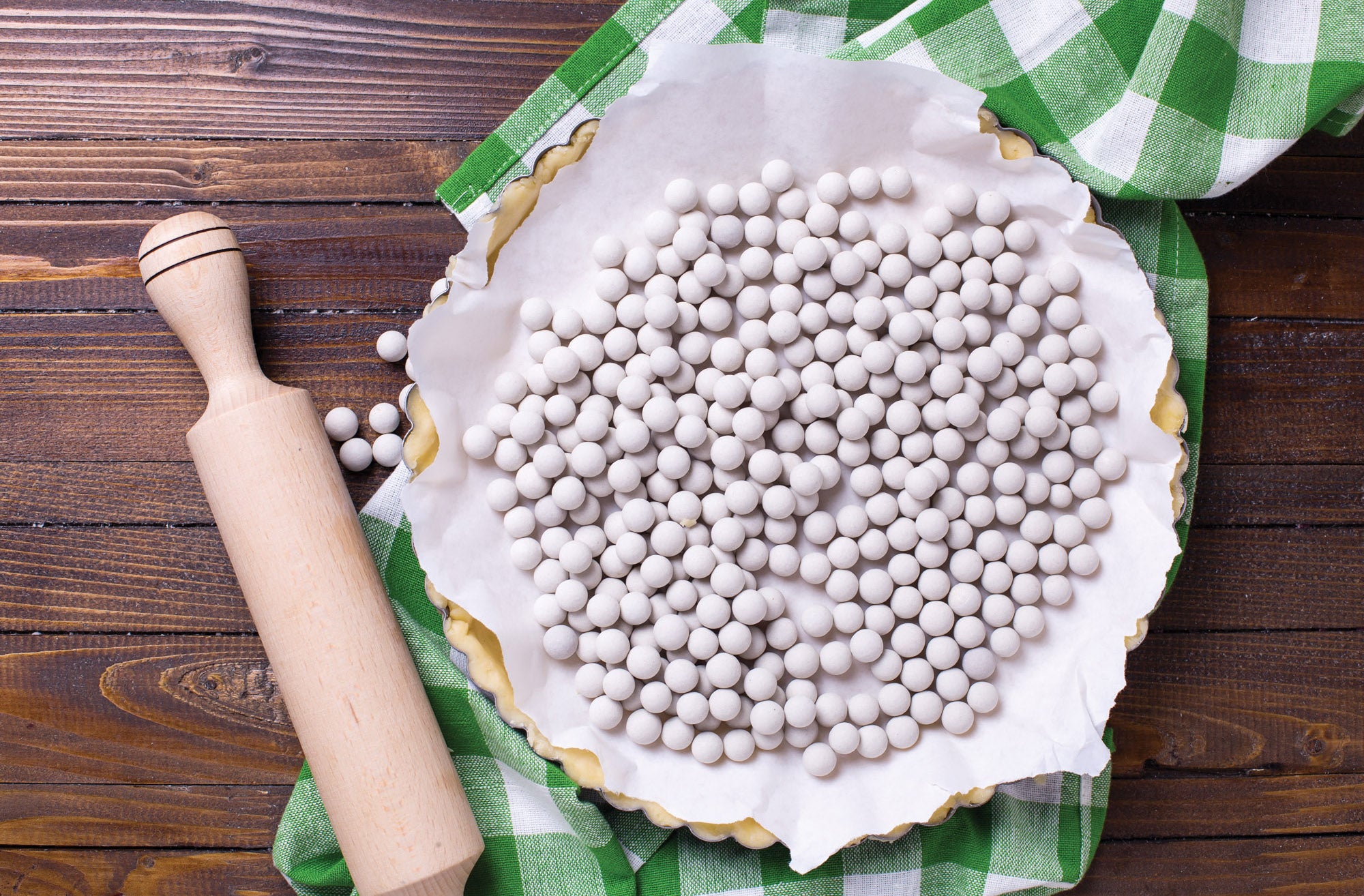 Mrs. Anderson's Baking Ceramic Pie Weights are used for baking a pie. The pie dish sits on a green and white towel, beside a wooden rolling pin.