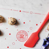 Red spatula and cookie scoop on a King Arthur Baking Company Cookie Mat with cookie dough balls.