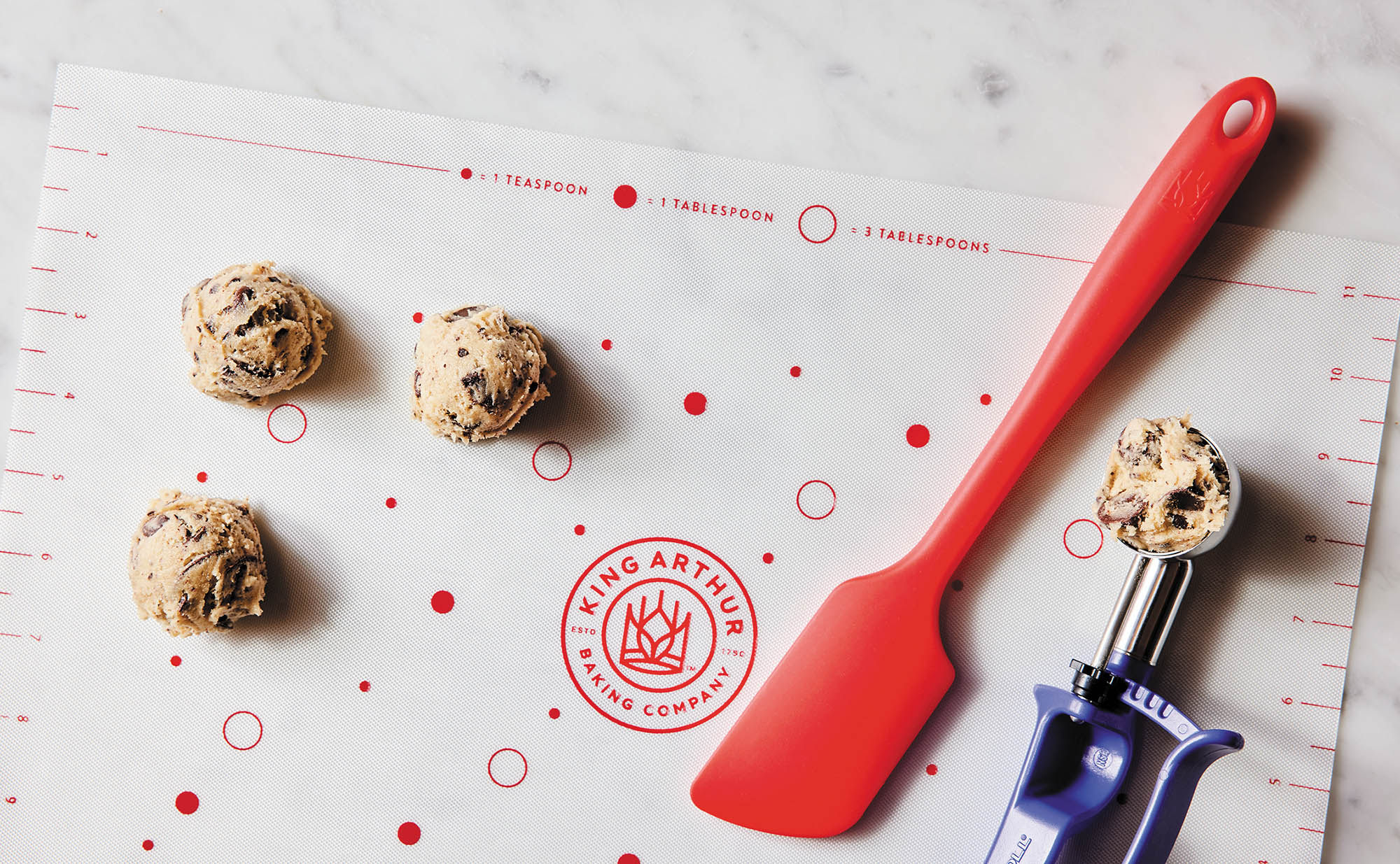Red spatula and cookie scoop on a King Arthur Baking Company Cookie Mat with cookie dough balls.