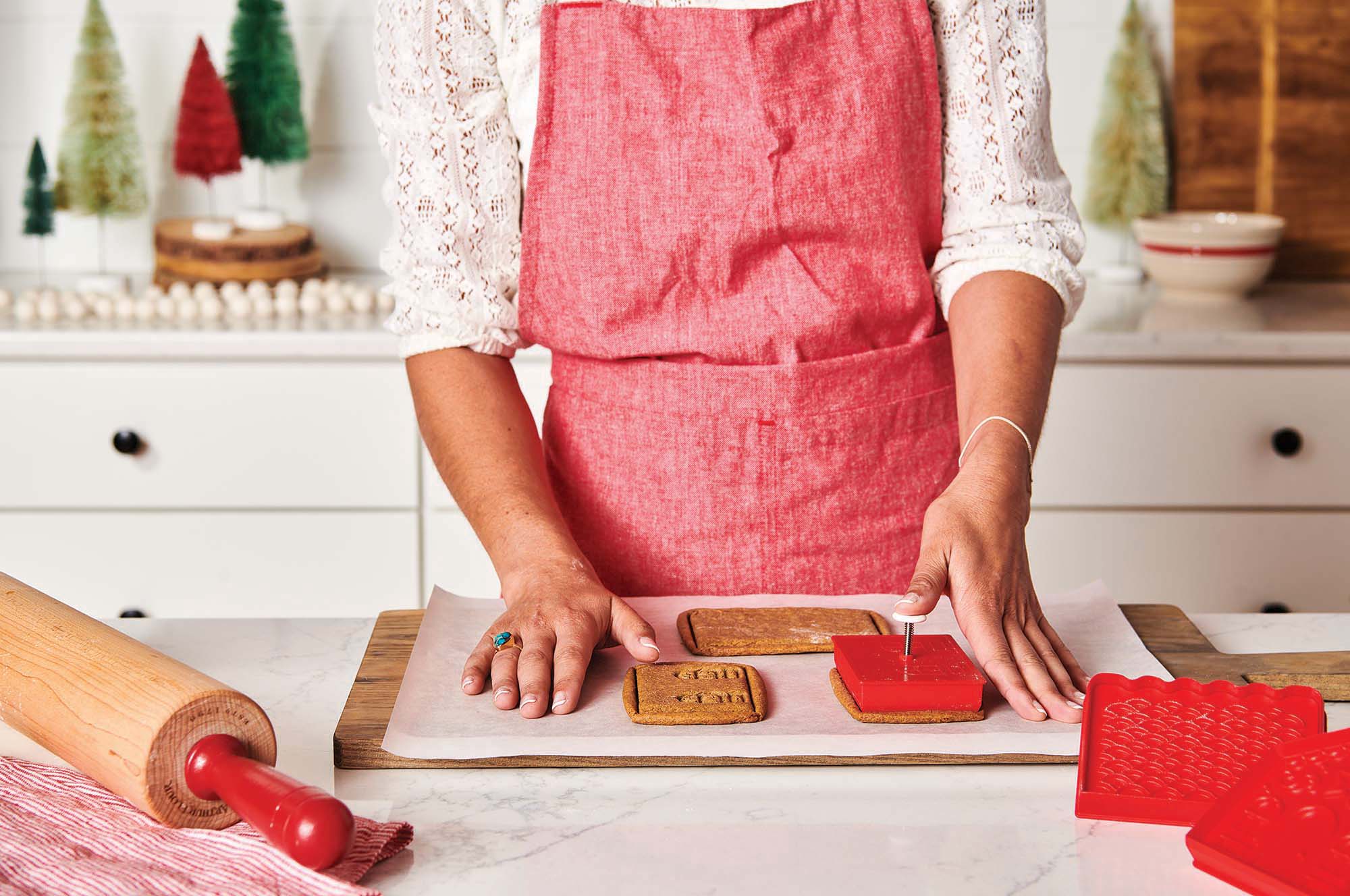 A person in a red apron uses the King Arthur Baking Company 3-D Gingerbread Cutter Set to make gingerbread houses in a kitchen.