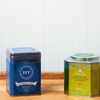 An assortment of Harney & Sons tea sits on a wooden countertop. The assortment includes the following teas: Blueberry Green and Green Tea with Coconut.