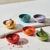 Six Le Creuset Pinch Bowls in assorted colors sit on a white marble countertop. Each bowl holds ingredients, including spices and herbs.