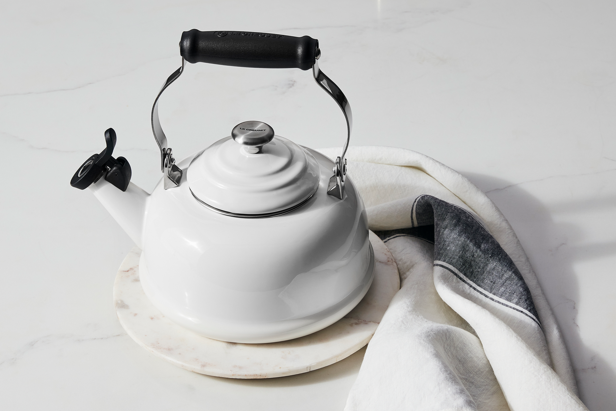 A Le Creuset Classic Kettle in White sits on a marble countertop alongside a kitchen towel.