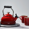 A Le Creuset Classic Kettle in Cherry sits on a marble countertop alongside a kitchen towel, two mugs, and flowers in a vase.