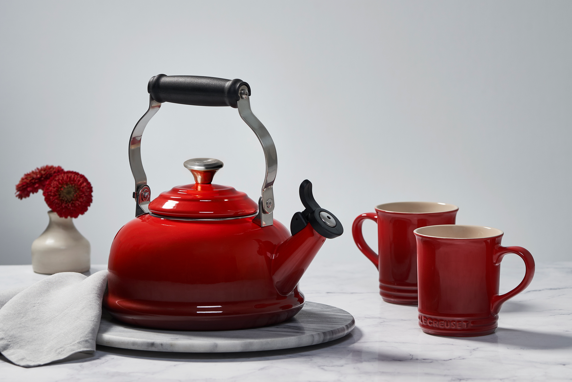 A Le Creuset Classic Kettle in Cherry sits on a marble countertop alongside a kitchen towel, two mugs, and flowers in a vase.