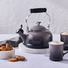 A Le Creuset Classic Kettle in Oyster sits on a marble countertop alongside a kitchen towel, two mugs, and a bowl of cookies.
