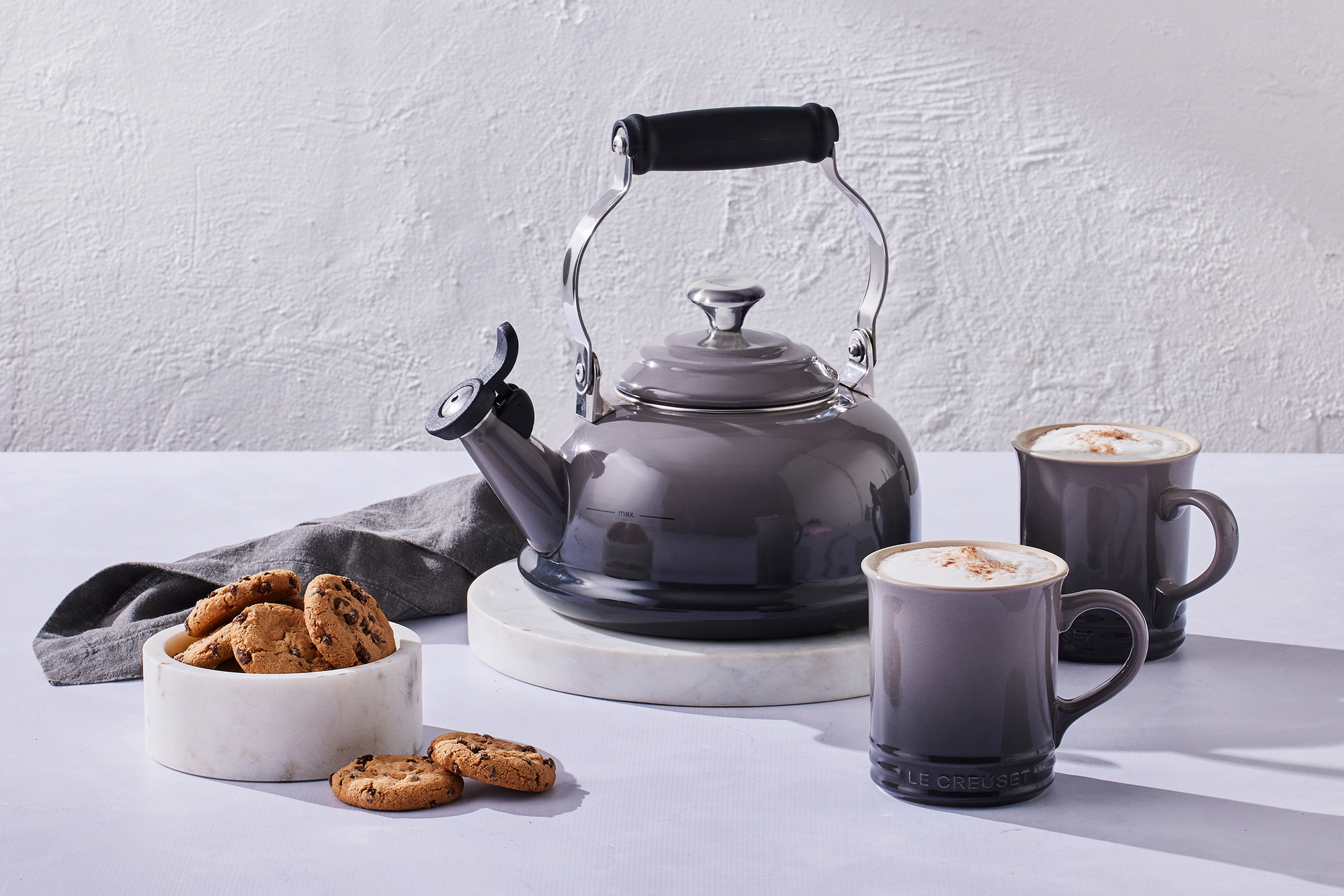 A Le Creuset Classic Kettle in Oyster sits on a marble countertop alongside a kitchen towel, two mugs, and a bowl of cookies.