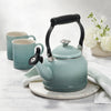 A Le Creuset Demi Kettle in Sea Salt sits on a marble countertop alongside two mugs, a kitchen towel, and potted flowers.