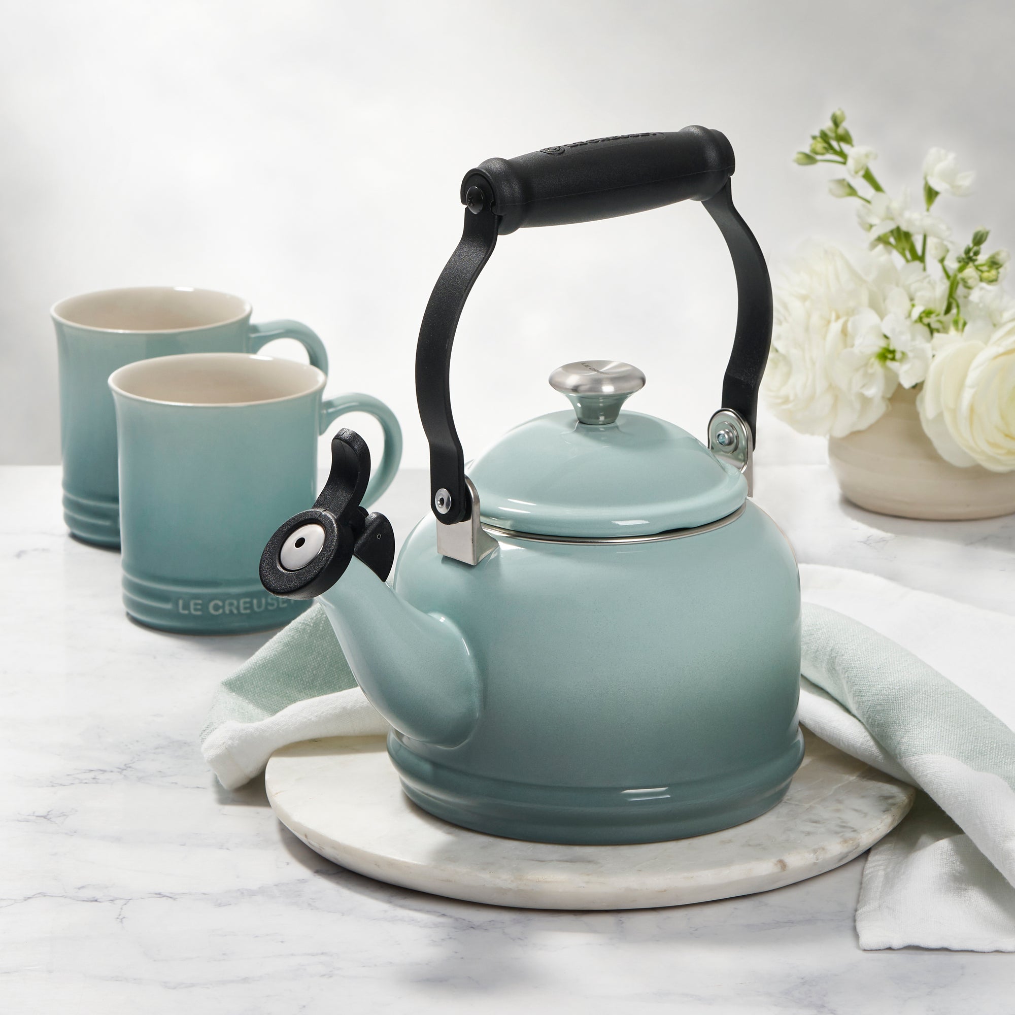 A Le Creuset Demi Kettle in Sea Salt sits on a marble countertop alongside two mugs, a kitchen towel, and potted flowers.